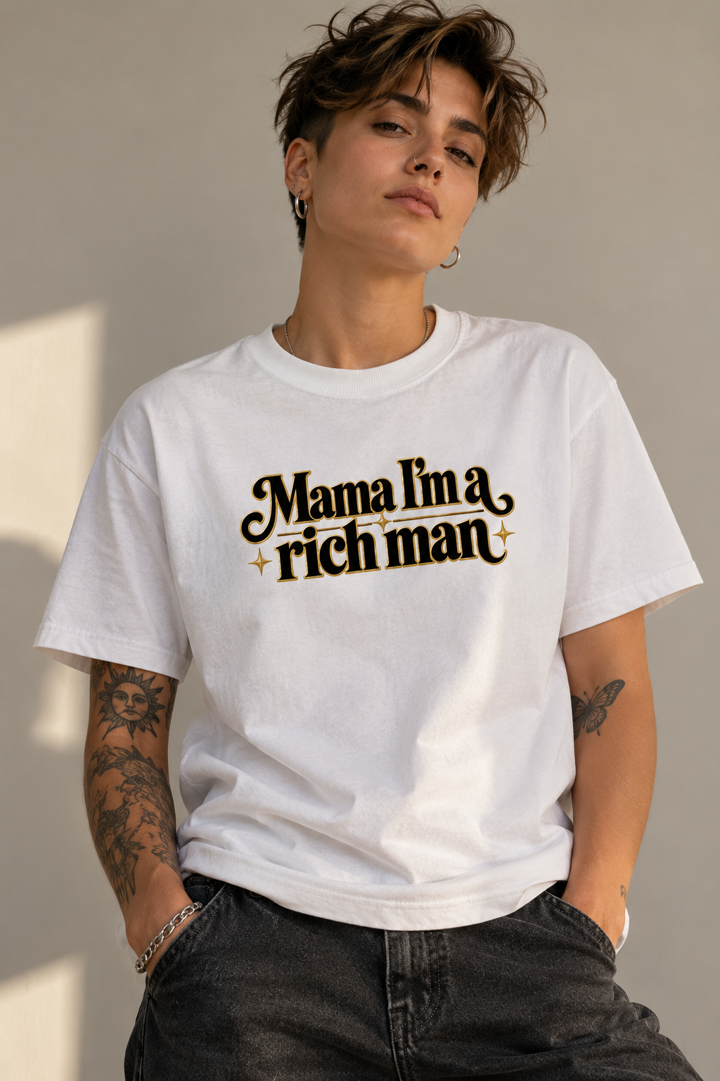 A person wearing a white graphic T-shirt with the phrase 'Mama I'm a rich man' printed in black and gold across the chest, paired with dark jeans against a neutral indoor wall.