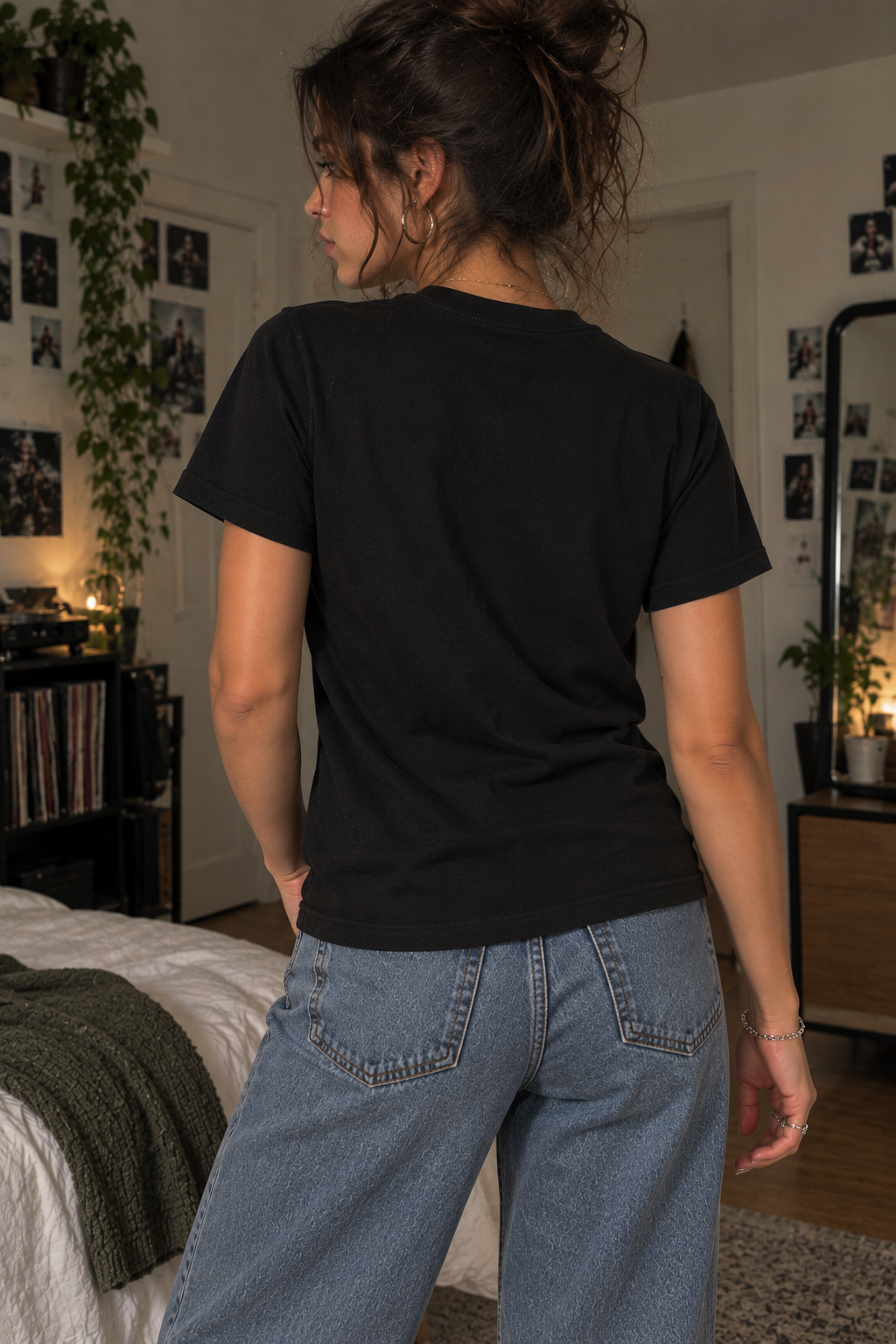 A woman wearing a plain black short-sleeve t-shirt and light blue jeans, photographed from behind in a cozy, dimly lit bedroom setting.