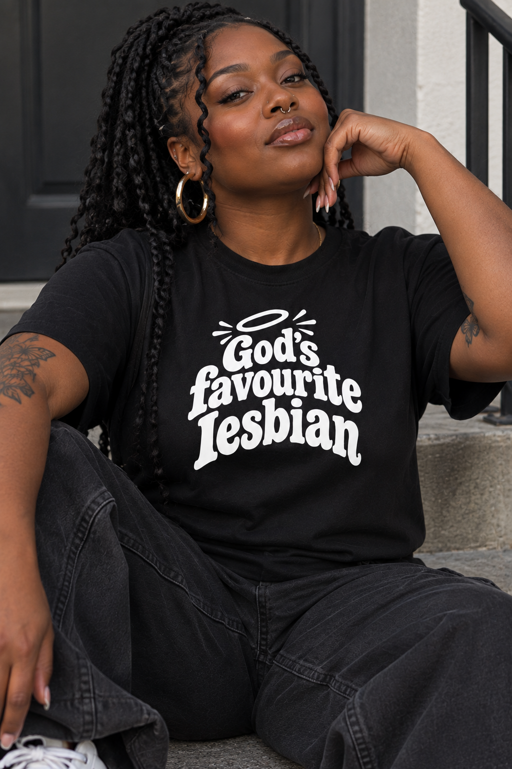 A woman sitting on outdoor steps wearing a black graphic t-shirt with the text 'God's favourite Lesbian' in white, paired with dark gray jeans.