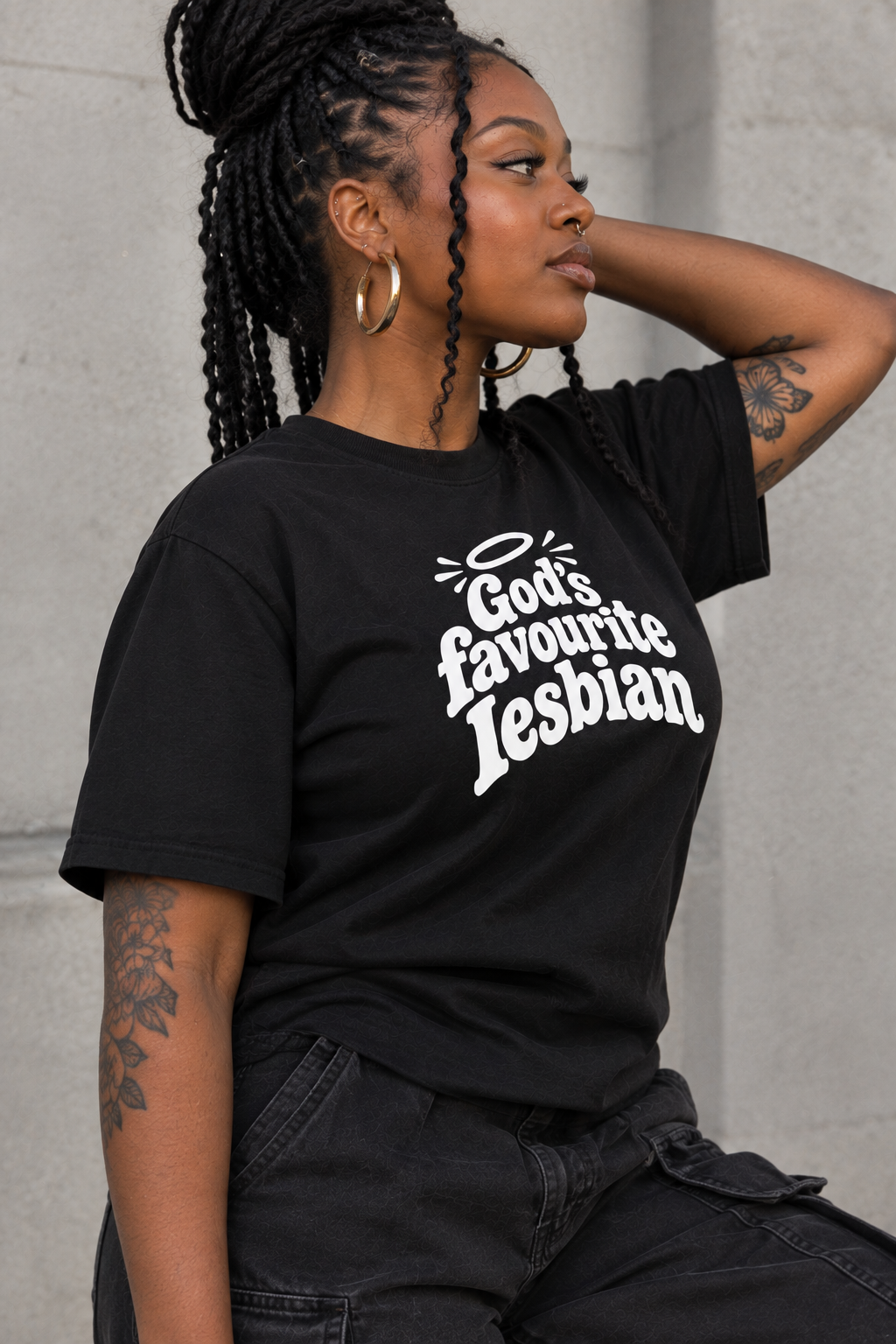 A woman wearing a black graphic t-shirt with the text 'God's favourite Lesbian' in white, posed against a gray concrete wall.