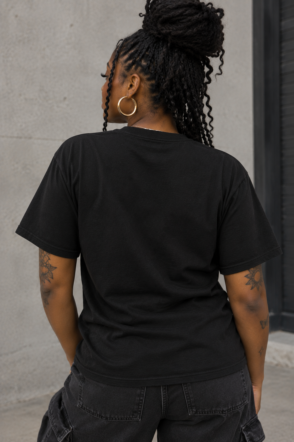 A woman with braided hair is shown from behind wearing a plain black short-sleeve t-shirt and dark cargo pants, standing against a concrete wall.