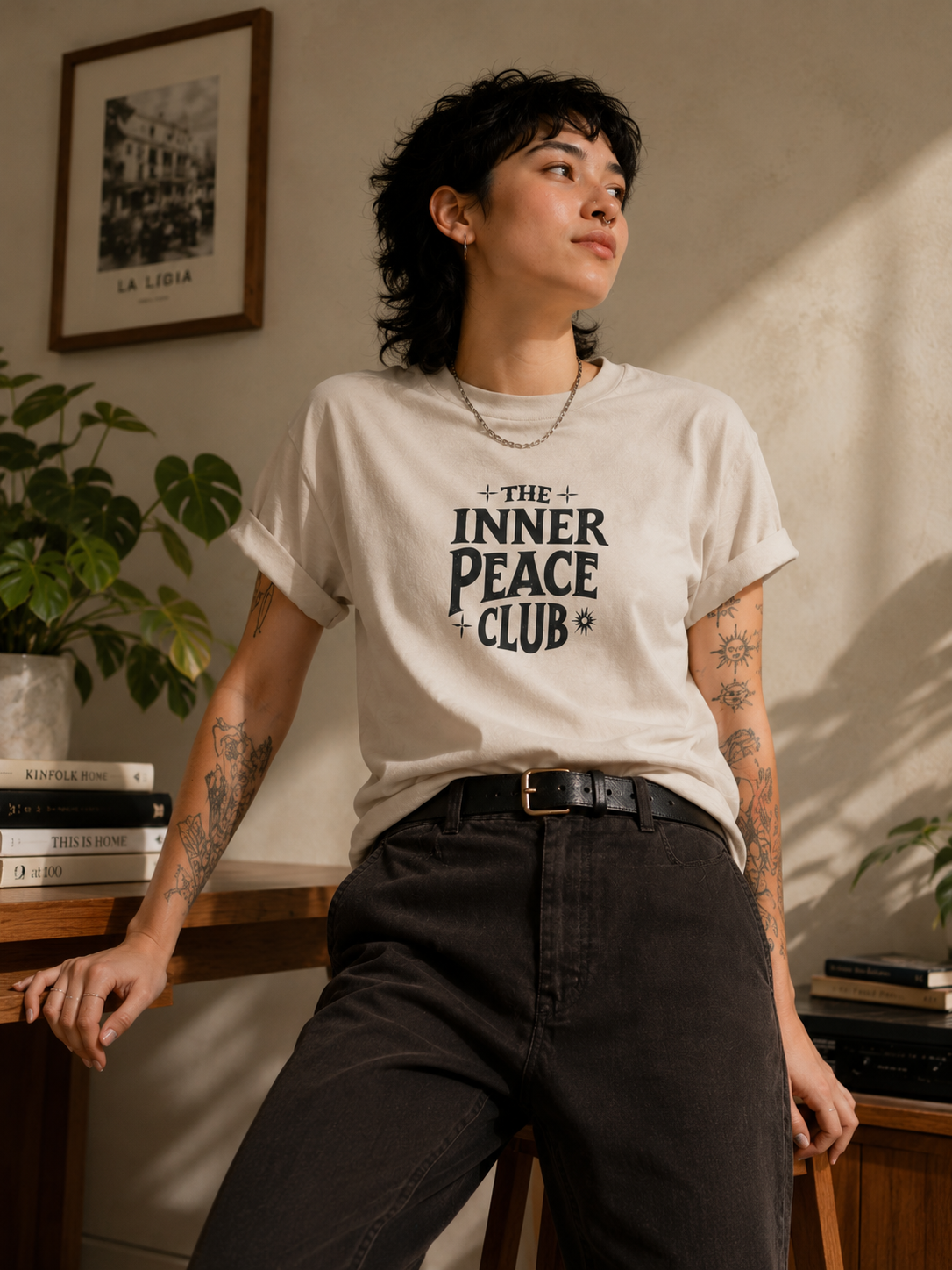 A person wearing a cream-colored graphic t-shirt with the text 'THE INNER PEACE CLUB' in black, paired with dark charcoal pants and a black belt, standing in a warmly lit room with a bookshelf and framed art in the background.