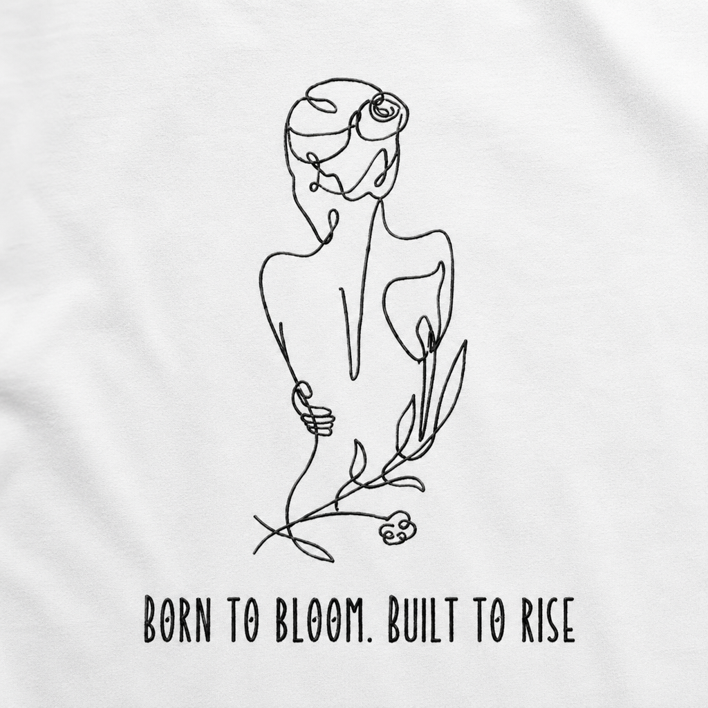 White t-shirt featuring a minimalist black line art illustration of a woman with flowers in her hair and the text 'BORN TO BLOOM. BUILT TO RISE' printed below.