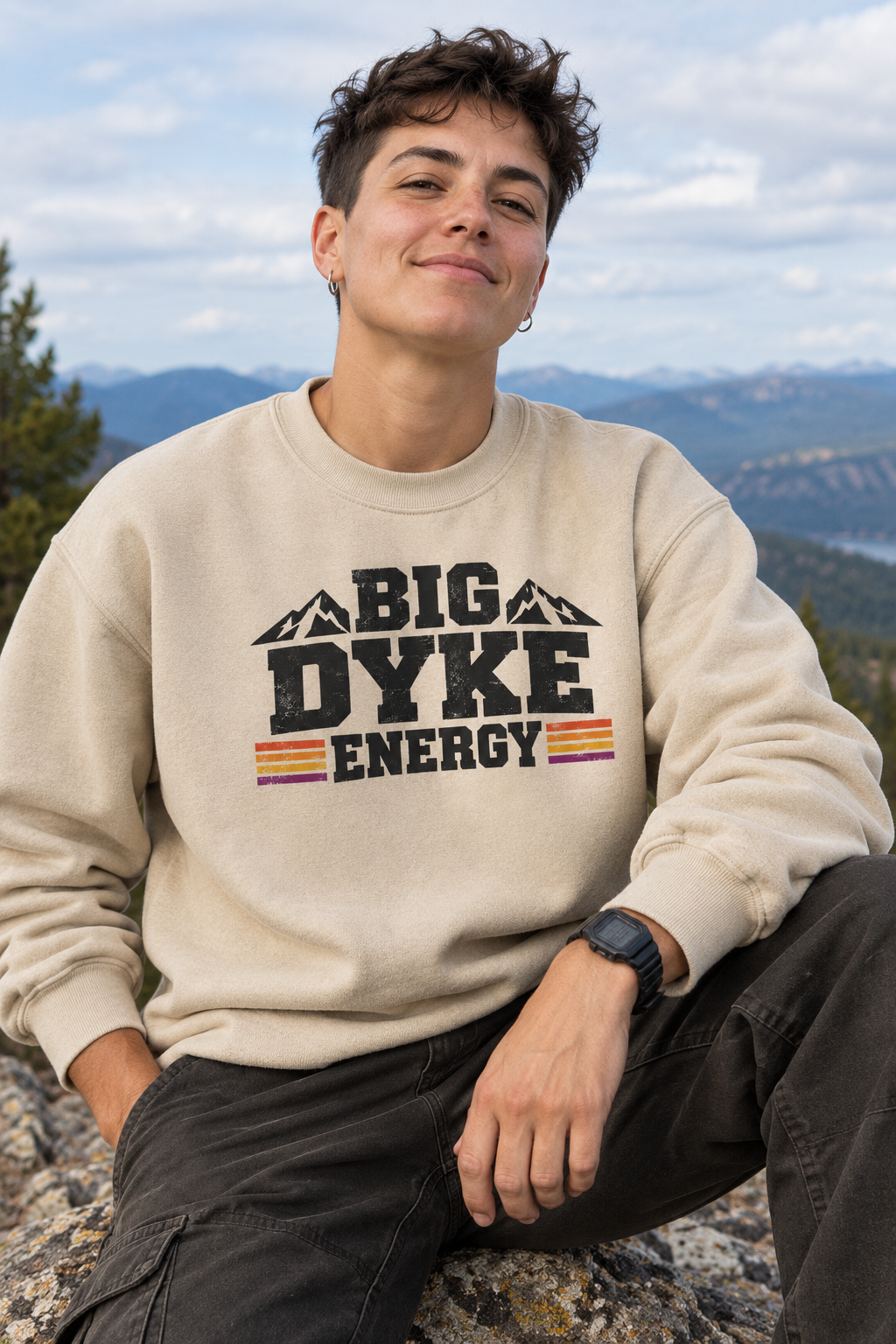 A man wearing a beige crewneck sweatshirt with a bold black graphic that reads 'BIG DYKE ENERGY' with mountain silhouettes and retro striped accents, sitting outdoors with a mountain landscape in the background.