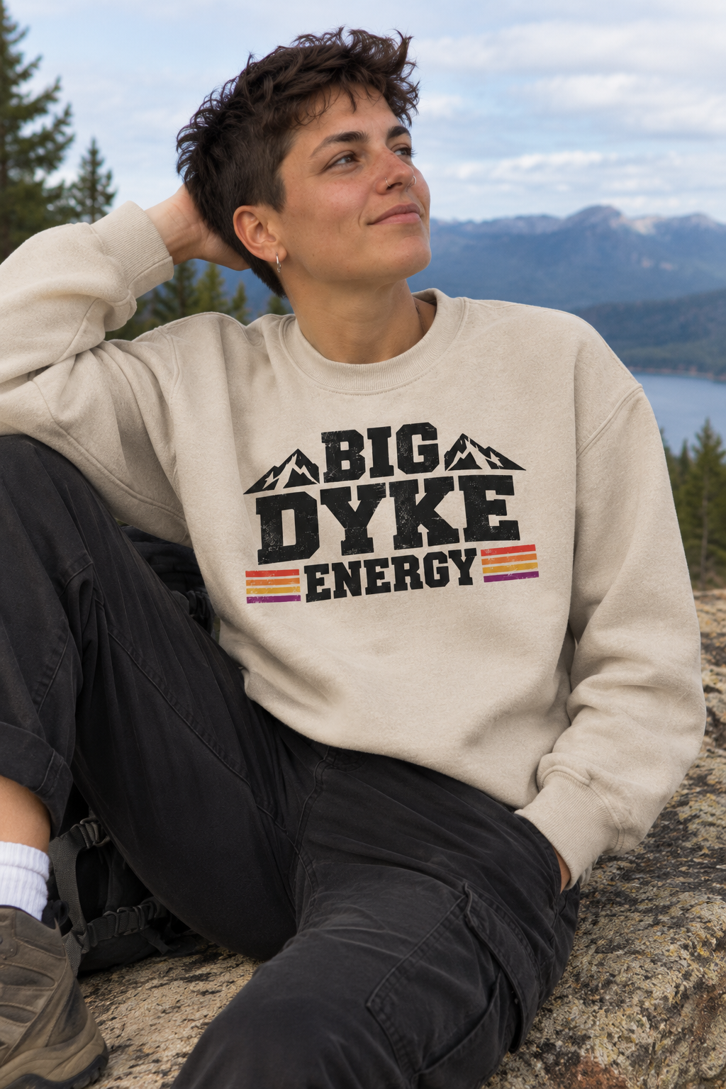A man wearing a beige crewneck sweatshirt with a bold black graphic that reads 'BIG DYKE ENERGY' with mountain silhouettes and red and yellow stripes, sitting outdoors on a rock with mountains and water in the background.