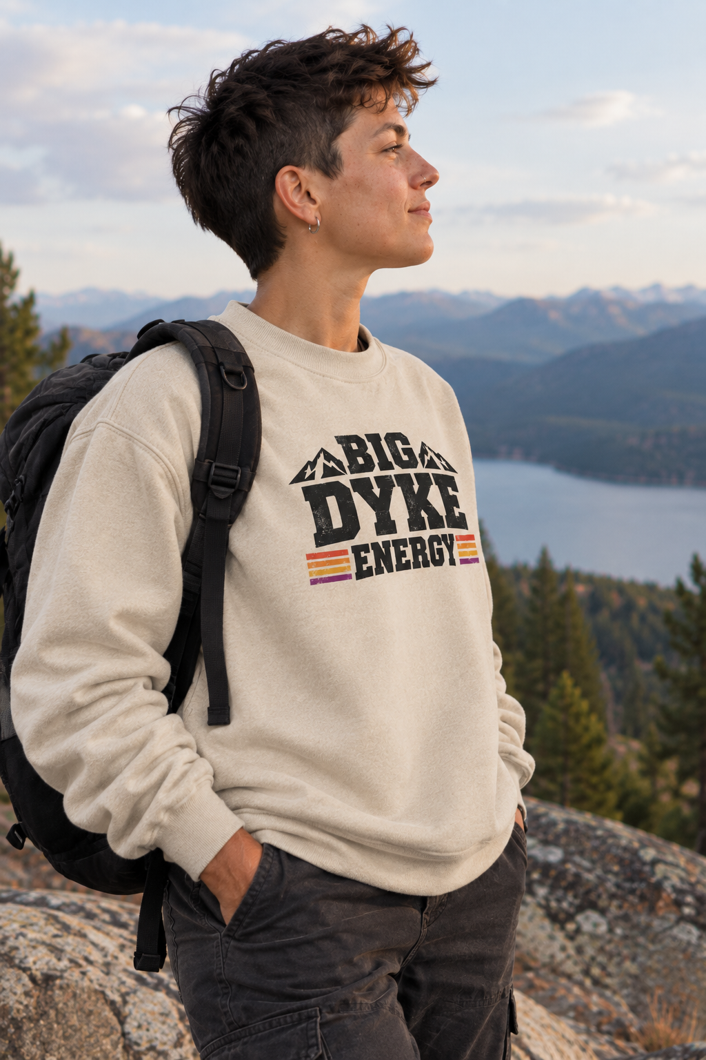 A man wearing a cream-colored sweatshirt with a 'BIG DYKE ENERGY' graphic stands on a rocky mountain peak with a lake and mountains in the background, carrying a black backpack.
