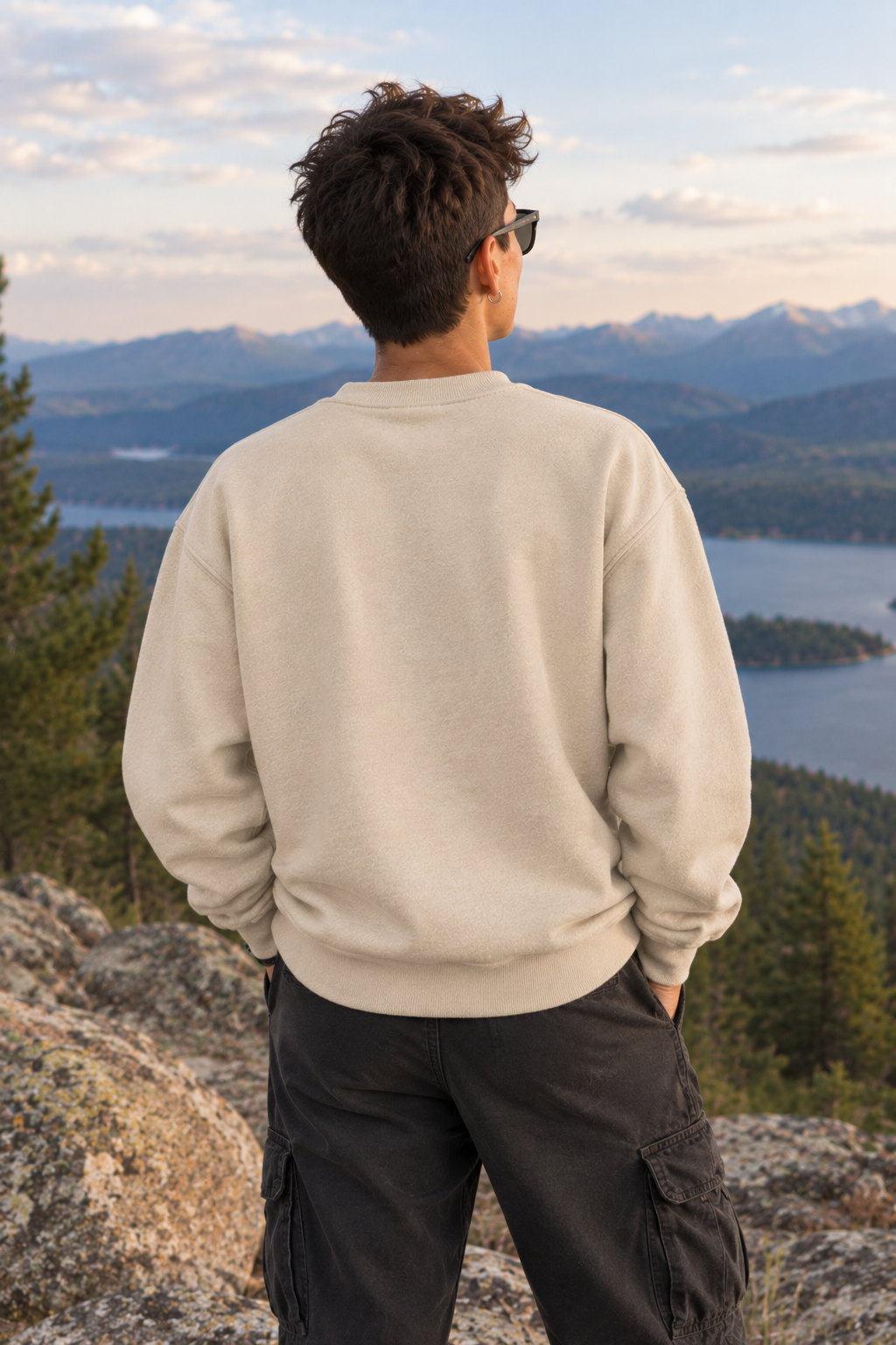 A man viewed from behind wearing a light beige crewneck sweatshirt and dark cargo pants, standing on a rocky mountain peak with a scenic lake and mountain range in the background.