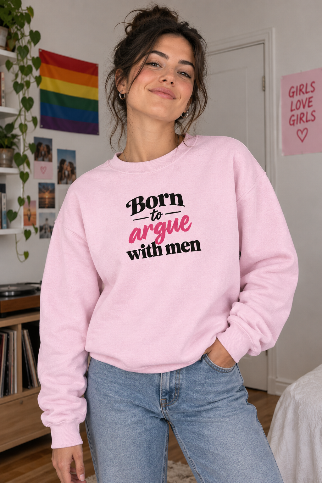 A woman wearing a light pink crewneck sweatshirt with the text 'Born to argue with men' in black and pink lettering, paired with light blue jeans, standing in a bedroom setting.