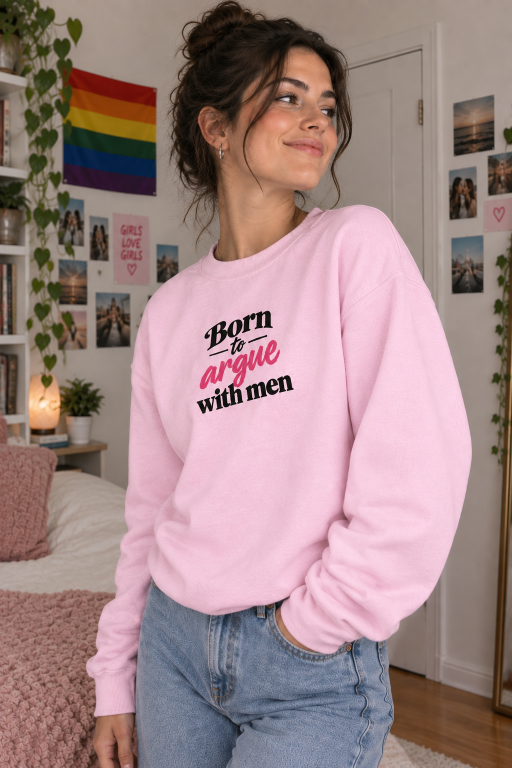 A woman wearing a light pink crewneck sweatshirt with the text 'Born to argue with men' in black and pink lettering, paired with light blue jeans in a bedroom setting.