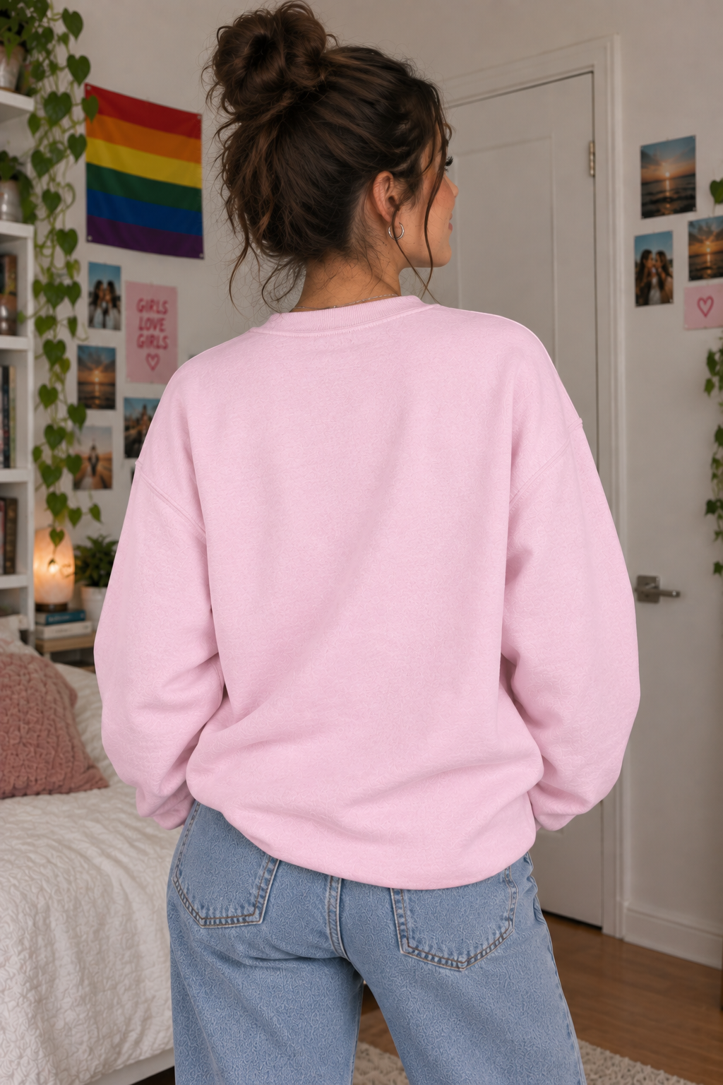 A woman with her hair in a bun is shown from behind wearing a light pink crewneck sweatshirt and light blue jeans in a bedroom setting.
