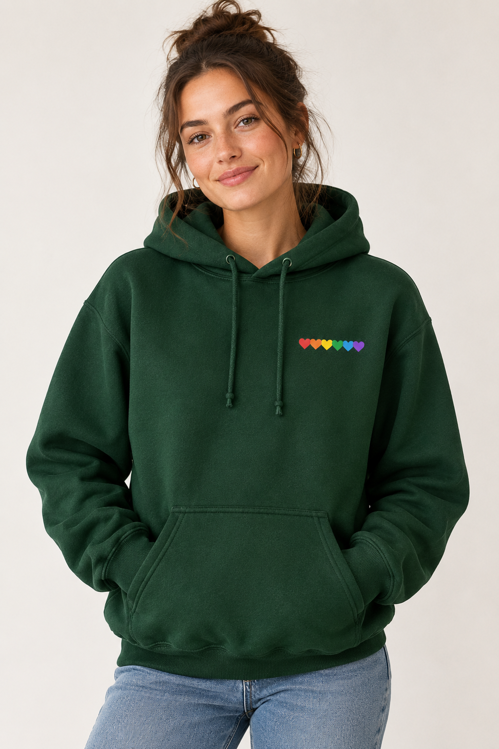 A woman wearing a dark green pullover hoodie with a small rainbow heart graphic on the left chest, paired with blue jeans, against a plain light background.