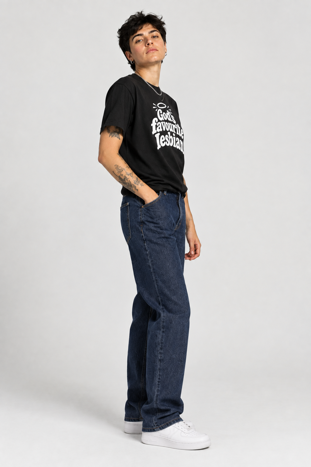 A person modeling a black graphic T-shirt with white text, paired with dark blue jeans and white sneakers, standing against a light gray studio background.