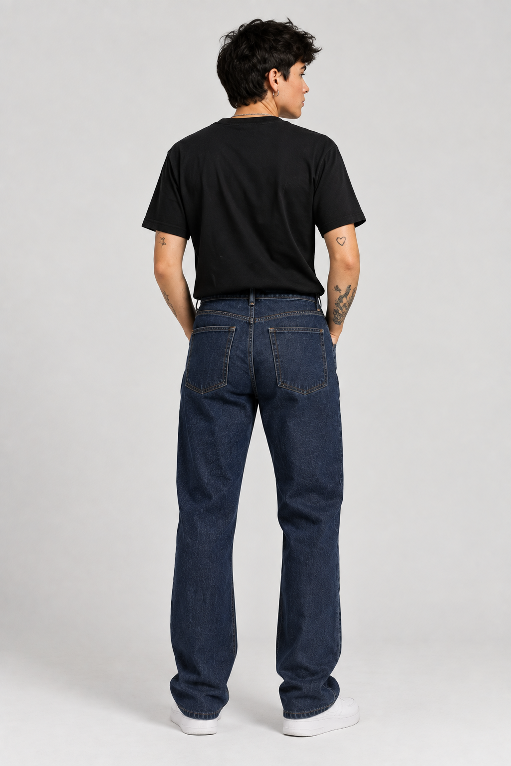 A man viewed from behind wearing a black t-shirt, dark blue straight-leg jeans, and white sneakers, standing against a light gray studio background.