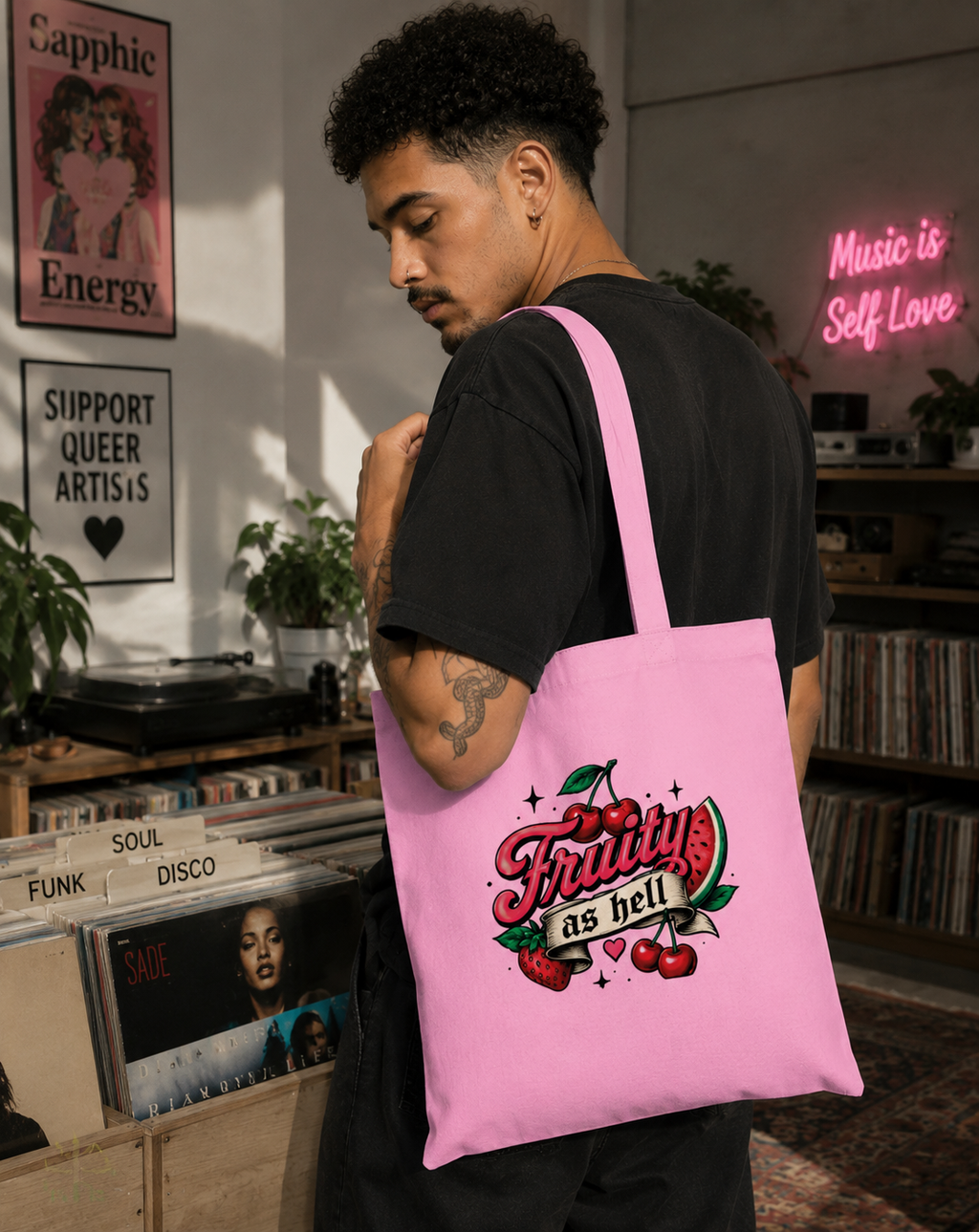 A man wearing a black t-shirt stands in a record store, carrying a pink canvas tote bag over his shoulder. The bag features a retro-style graphic with the text 'Fruity as hell' in red and white, decorated with cherries and green leaves.