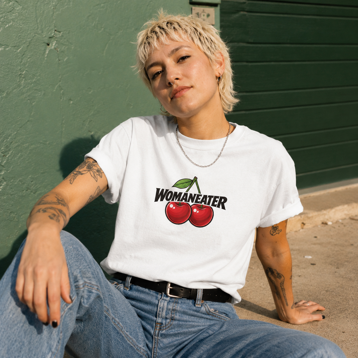 A person with short blonde hair sits outdoors against a green wall wearing a white graphic T-shirt with the word 'WOMANEATER' and two red cherries, paired with blue jeans.