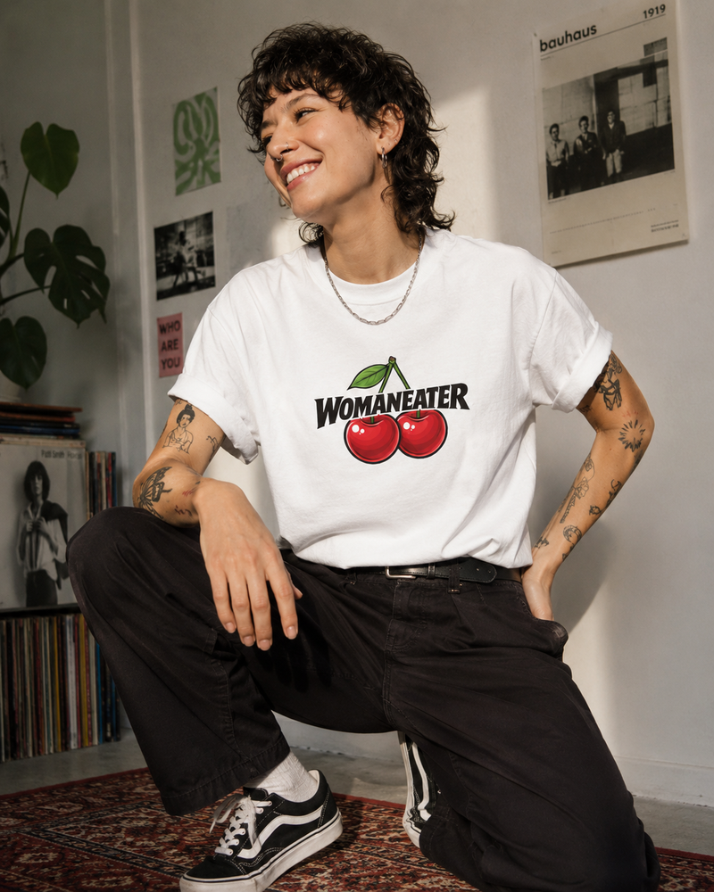 A person crouching indoors wearing a white graphic T-shirt with a red cherry illustration and the word 'WOMANEATER' across the chest, paired with black pants and black-and-white sneakers.