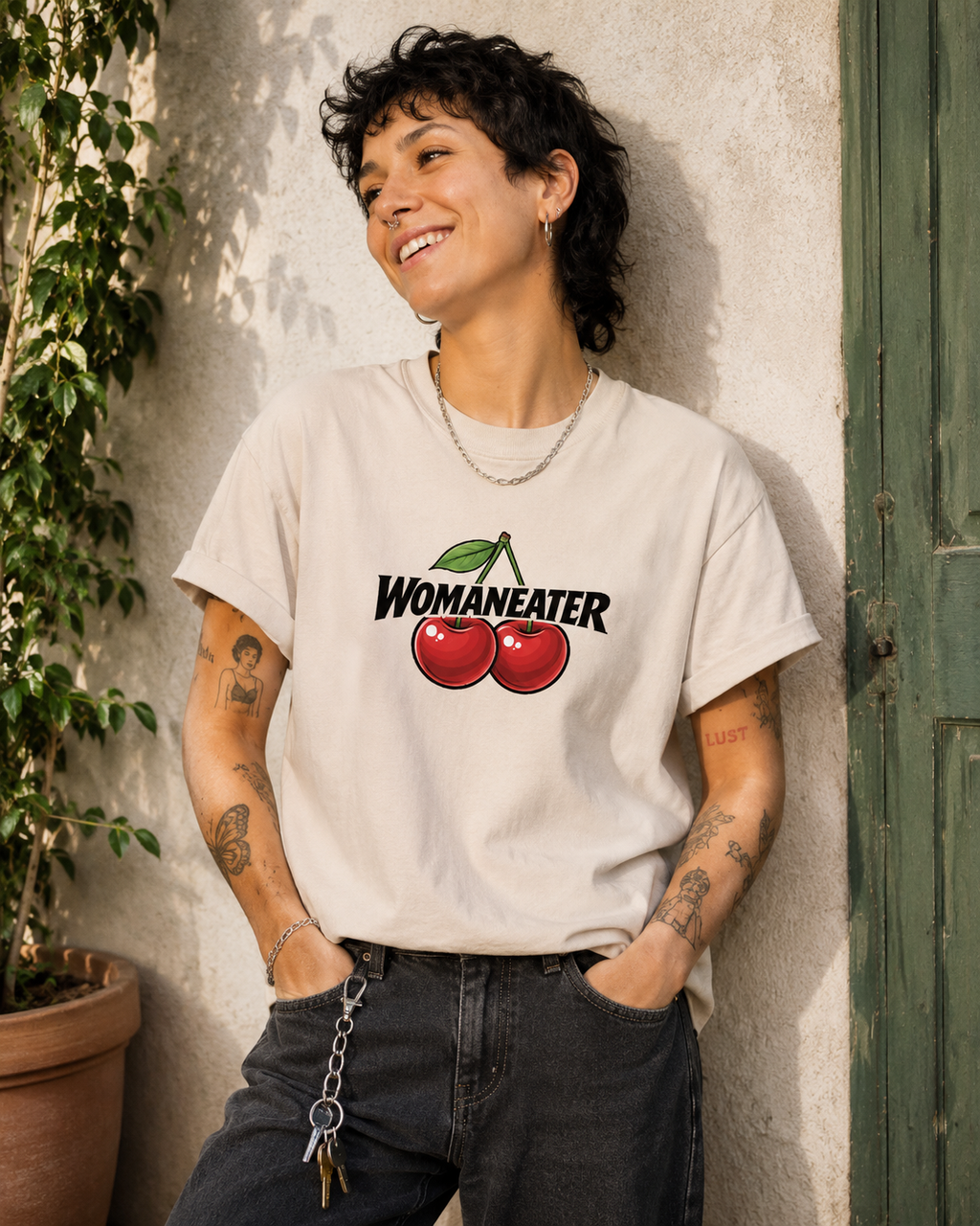 A person wearing a cream-colored graphic t-shirt with a cherry design and the text 'WOMANEATER' printed on the front, paired with dark jeans, leaning against a textured wall next to a potted plant.