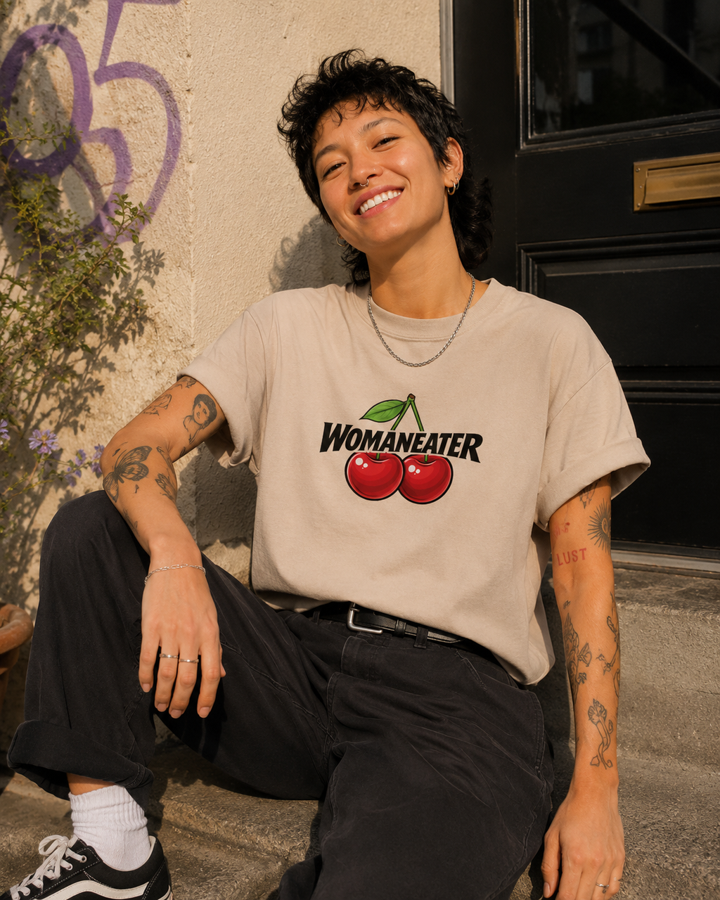 A smiling person with short dark hair sits outdoors against a wall and door, wearing a light beige graphic T-shirt with the word 'WOMANEATER' and two red cherries on the front, paired with dark pants and black-and-white sneakers.