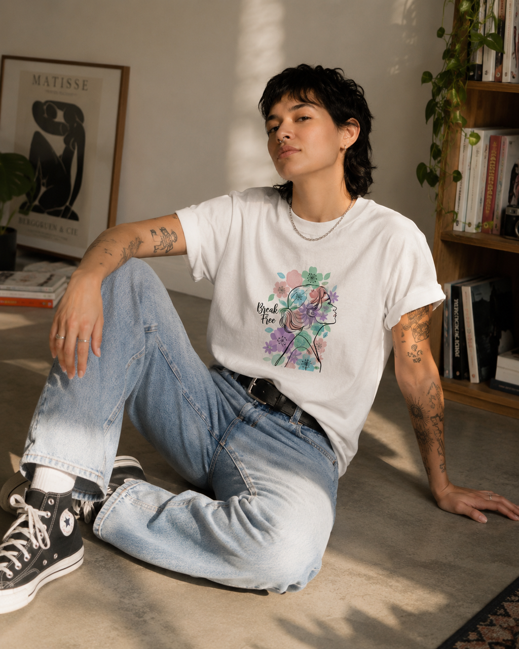 A person sitting on a concrete floor in a sunlit room, wearing a white graphic T-shirt with a colorful floral design, light-wash jeans, and black high-top sneakers.