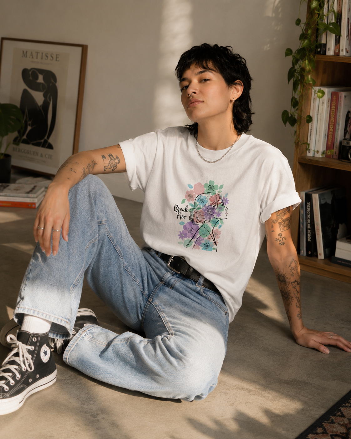 A person sitting on a concrete floor in a sunlit room, wearing a white graphic T-shirt with a colorful floral design, light-wash jeans, and black high-top sneakers.