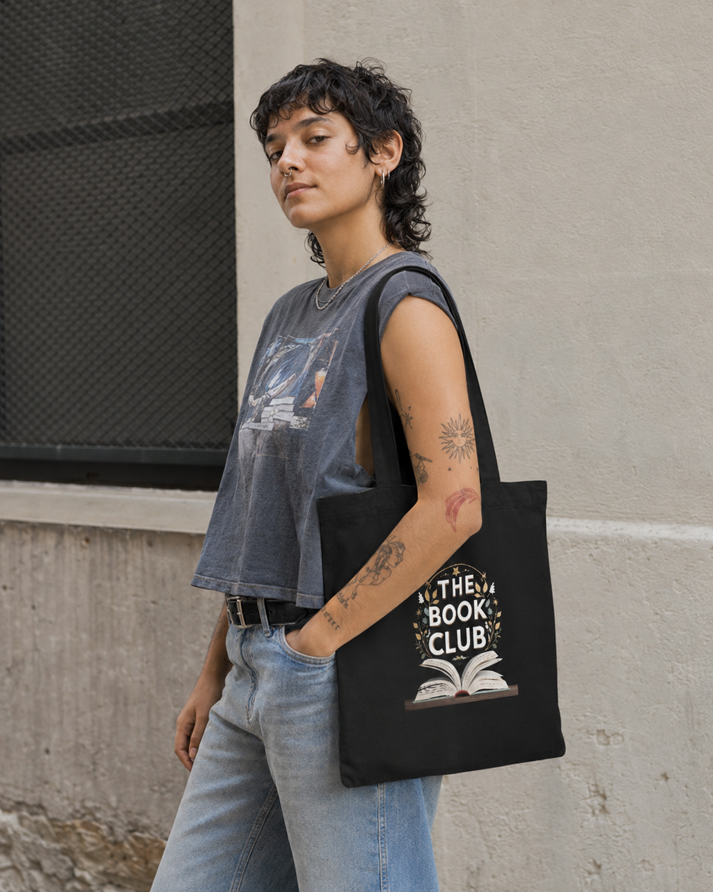 A person wearing a gray sleeveless top and light blue jeans carries a black tote bag with a white graphic that reads 'IN THE BOOK CLUB' above an open book illustration, standing against a concrete wall.