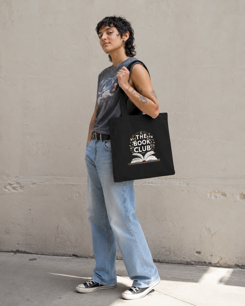 A person standing against a textured concrete wall, wearing a gray sleeveless graphic top, light blue jeans, and black sneakers, while carrying a black tote bag with a white 'The Best Book Club' graphic.