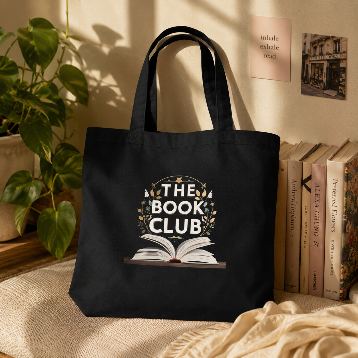 A black canvas tote bag with a 'THE BOOK CLUB' graphic printed on the front, styled on a beige fabric surface next to books, a potted plant, and a cream throw blanket.
