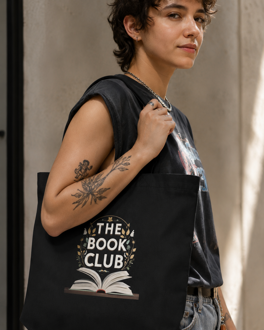 A person wearing a black sleeveless shirt and jeans carries a black tote bag printed with the words 'THE BOOK CLUB' and an open book graphic, photographed against a textured concrete wall.