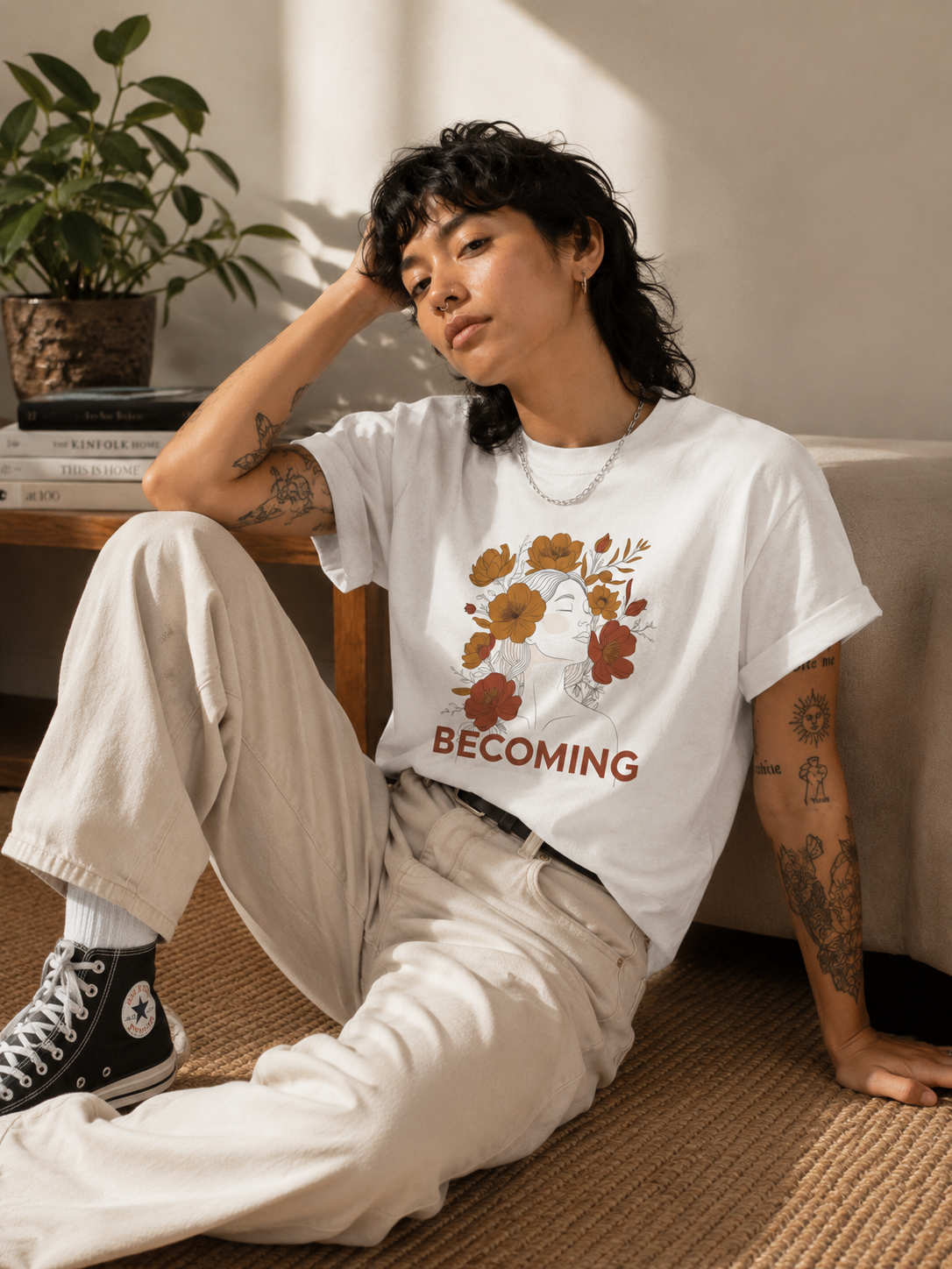 A person sitting indoors wearing a white graphic T-shirt with a floral print and the word 'BECOMING' in red, paired with light beige pants and black high-top sneakers.
