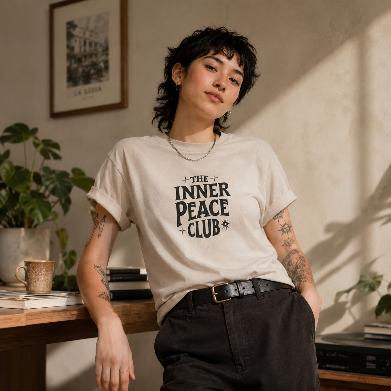 A person wearing a cream-colored graphic t-shirt with the text 'THE INNER PEACE CLUB' printed in black, paired with black pants, standing in a warmly lit room with plants and a framed picture in the background.