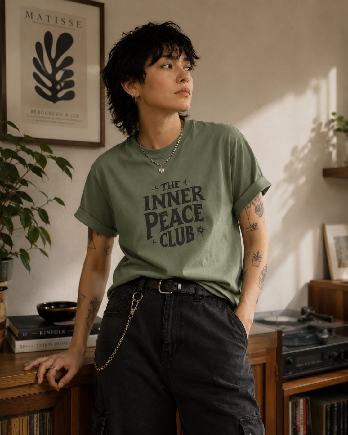 A person standing indoors wearing an olive green graphic T-shirt with the words 'THE INNER PEACE CLUB' printed in dark lettering, paired with dark pants and jewelry, in a warm, book-filled room.