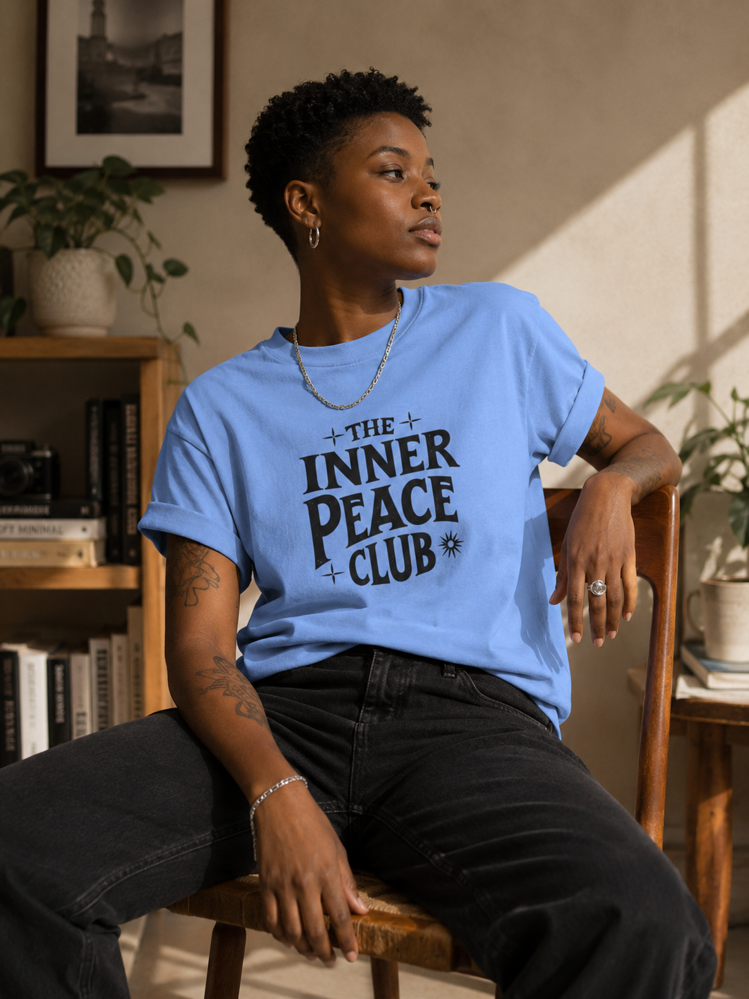 A person sitting on a wooden chair wearing a light blue graphic T-shirt with the text 'THE INNER PEACE CLUB' in dark lettering, paired with dark jeans in a warmly lit indoor room.