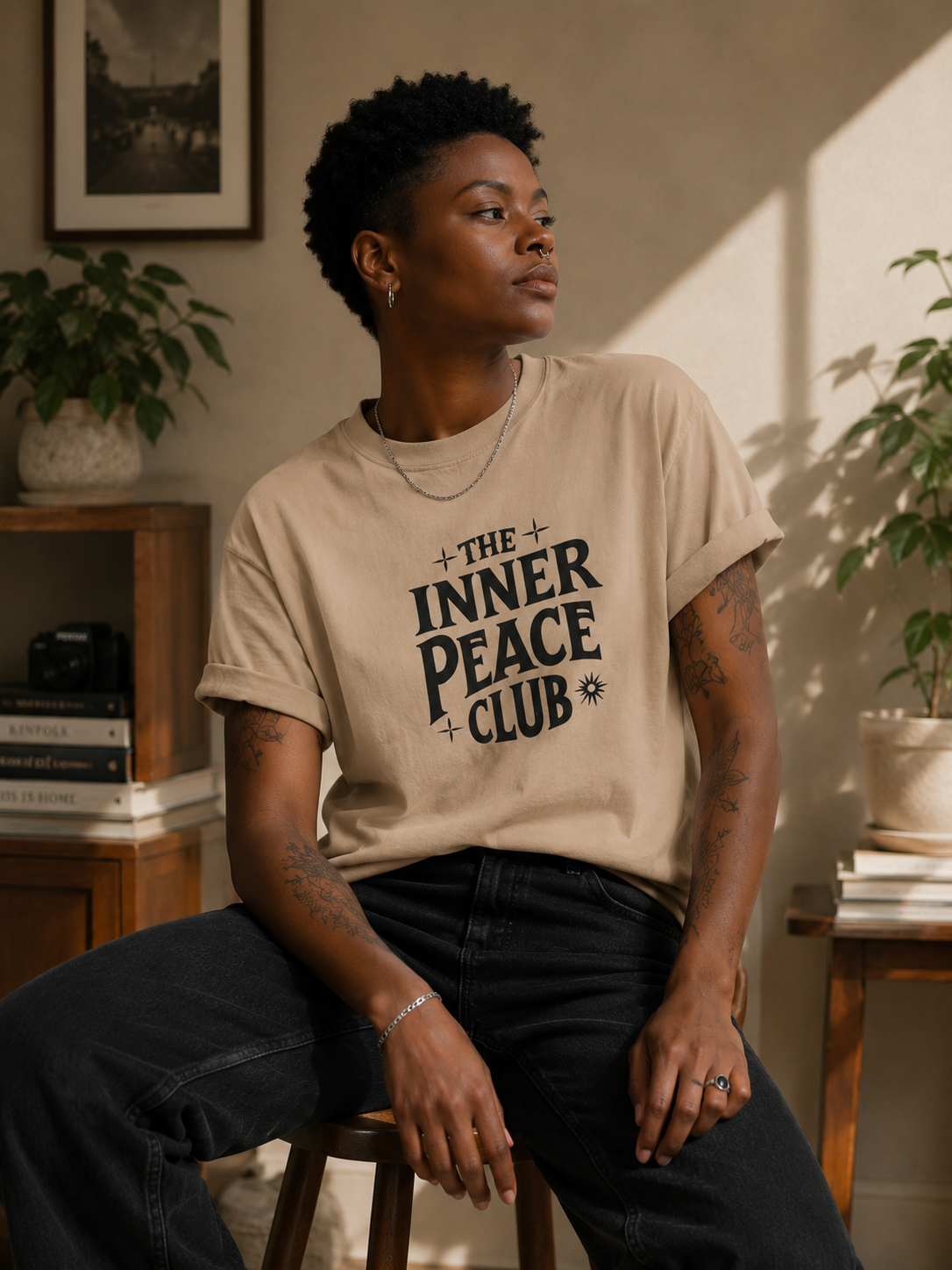 A person seated indoors wearing a beige graphic T-shirt with the words “THE INNER PEACE CLUB” in dark lettering, paired with dark jeans. The room has warm lighting, plants, books, and wooden furniture in the background.