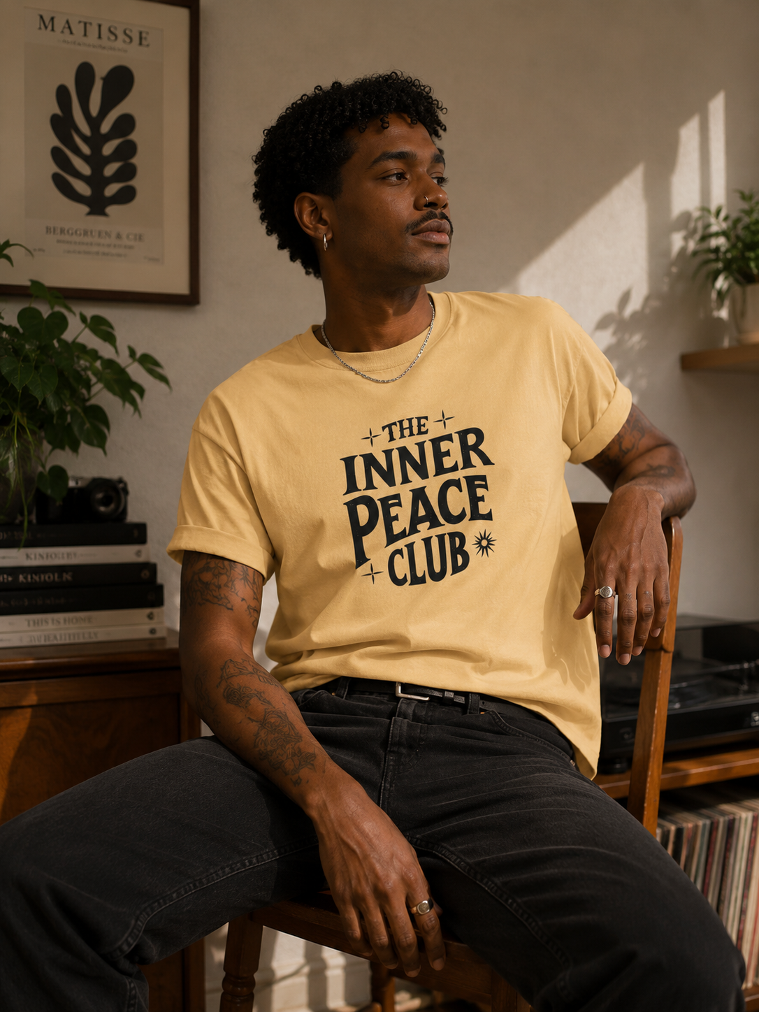 A man sitting on a wooden chair wearing a pale yellow graphic t-shirt with the text 'THE INNER PEACE CLUB' in black. The setting is a warm, sunlit room with a framed botanical print on the wall, plants, and vinyl records in the background.