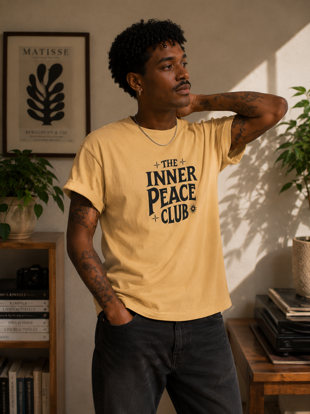 A man wearing a beige graphic t-shirt with the text 'THE INNER PEACE CLUB' printed in black, standing in a warmly lit room with plants and books in the background.