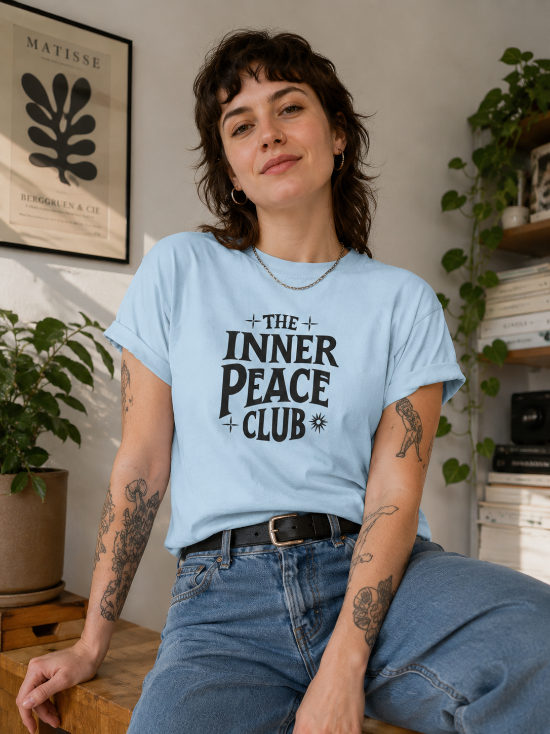 A woman wearing a light blue graphic t-shirt with the text 'THE INNER PEACE CLUB' printed in black, sitting in a cozy room with plants and books in the background.