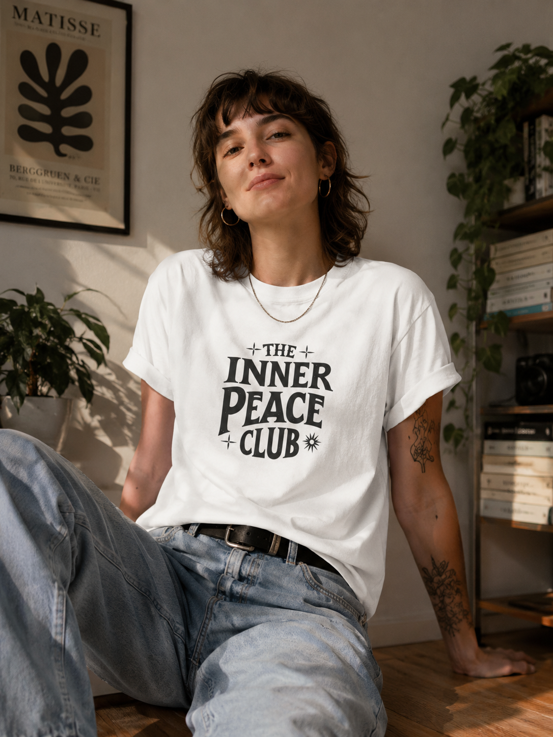 A woman wearing a white graphic t-shirt with the text 'THE INNER PEACE CLUB' in black, paired with light-wash blue jeans, sitting in a cozy, sunlit room with plants and a bookshelf.