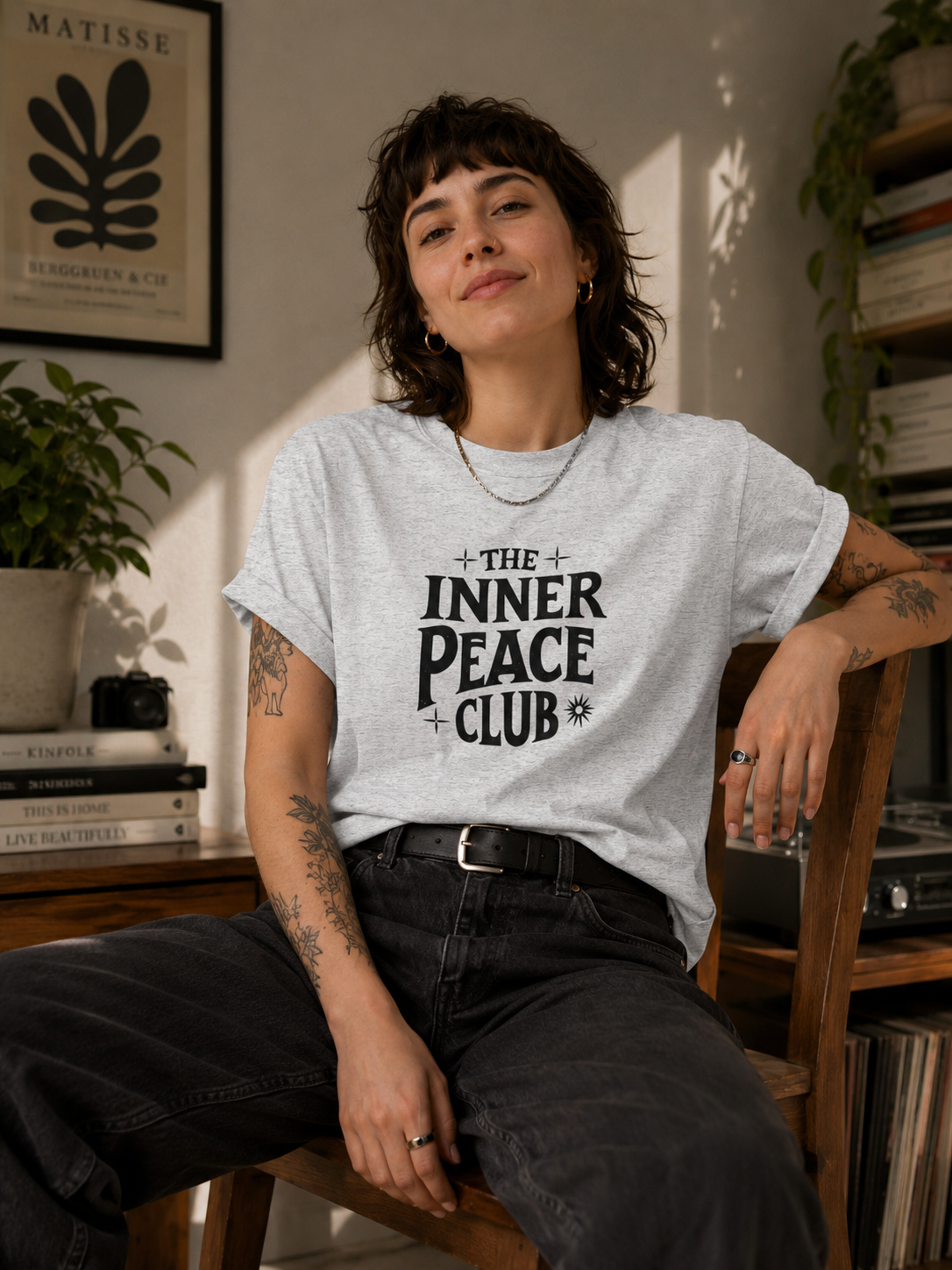 A woman with short dark hair sits on a wooden chair in a sunlit room, wearing a light heather gray graphic T-shirt with the text 'THE INNER PEACE CLUB' in black. She is also wearing dark jeans and a black belt.