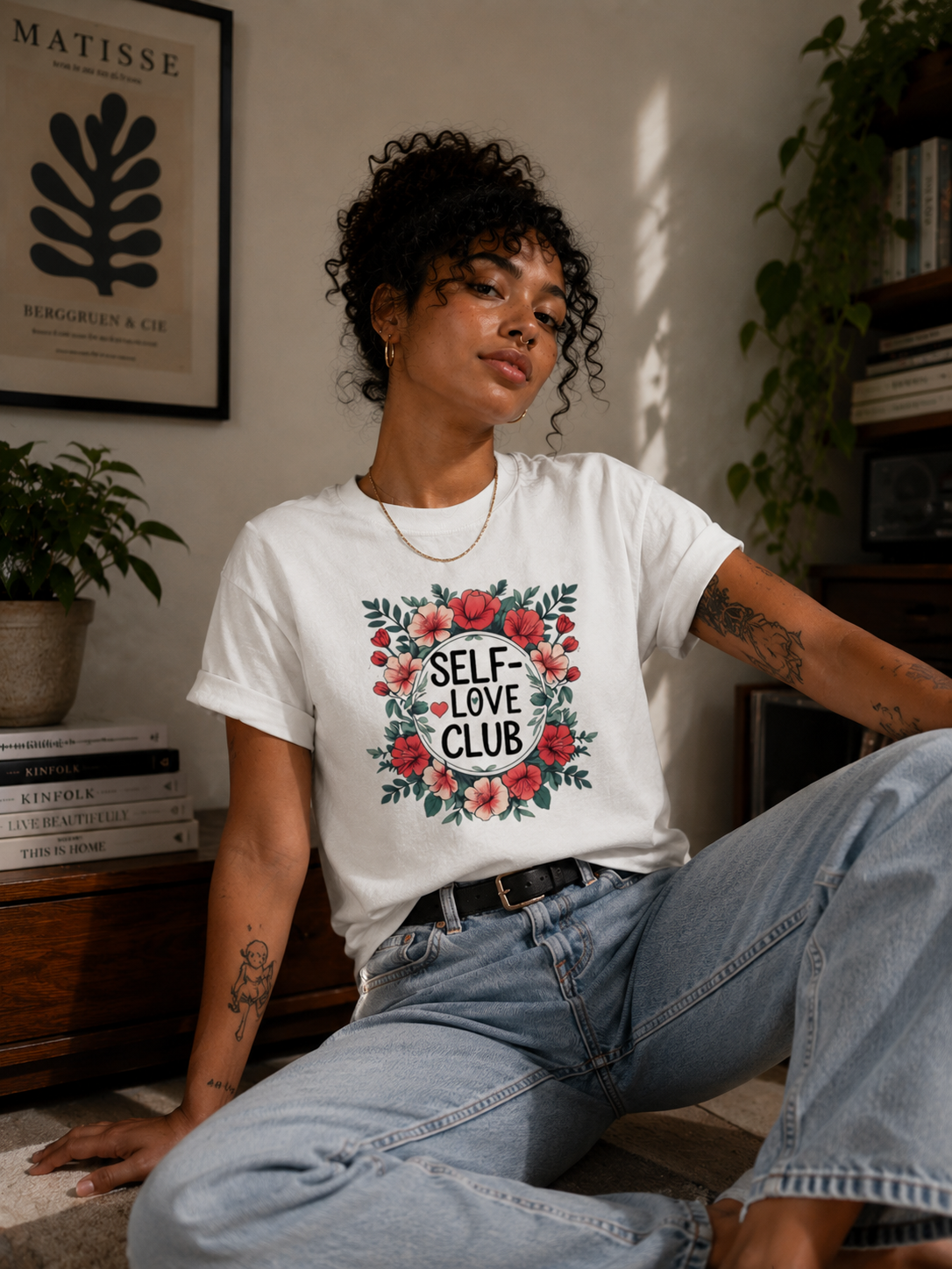 A woman wearing a white graphic t-shirt with a red and green floral design that reads 'SELF-LOVE CLUB' in black text, paired with light blue jeans, sitting in a cozy indoor setting.