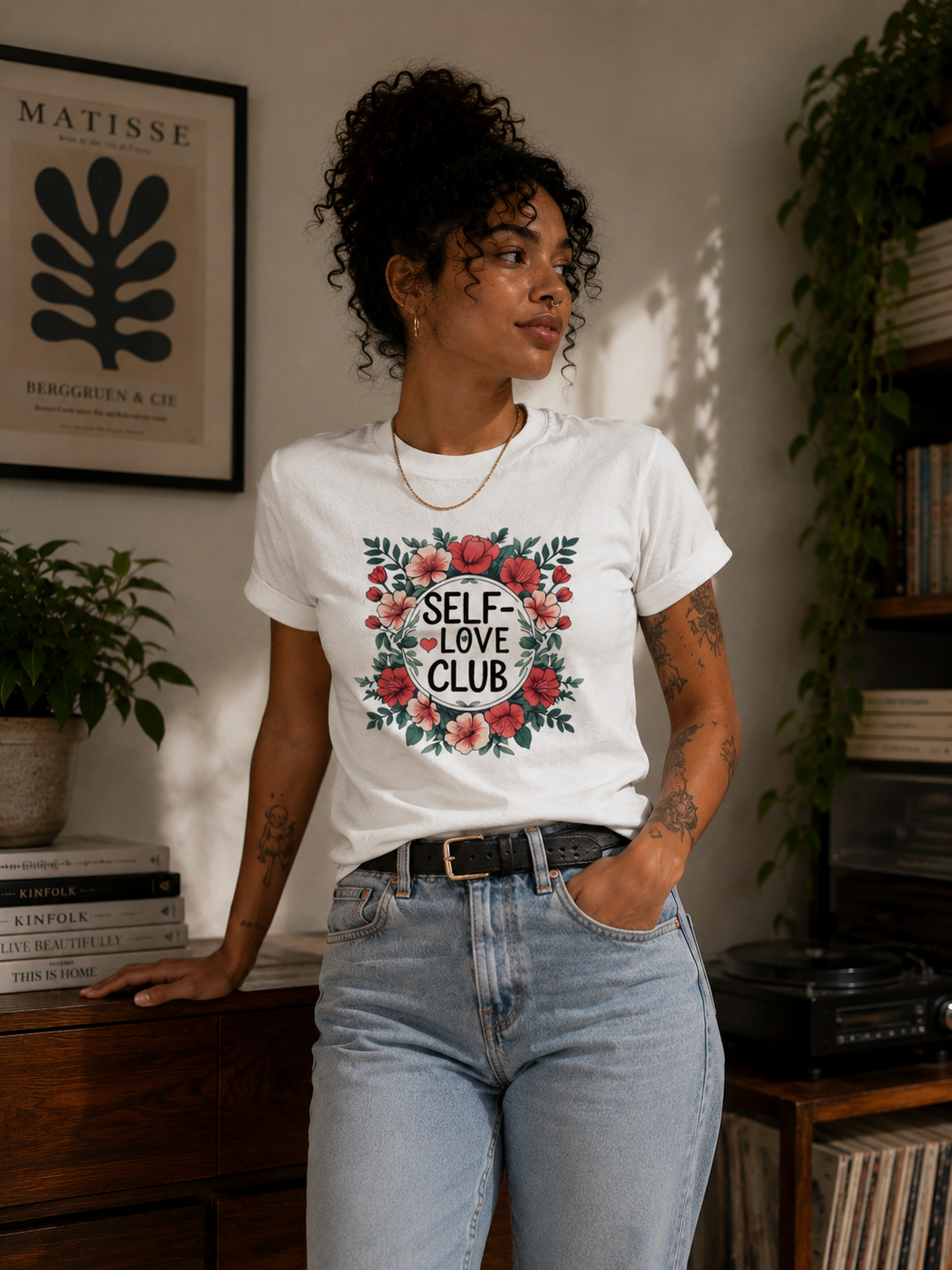 A woman wearing a white graphic t-shirt with a floral design and the text 'SELF-LOVE CLUB' paired with light-wash blue jeans, standing in a cozy, sunlit room with books and plants.