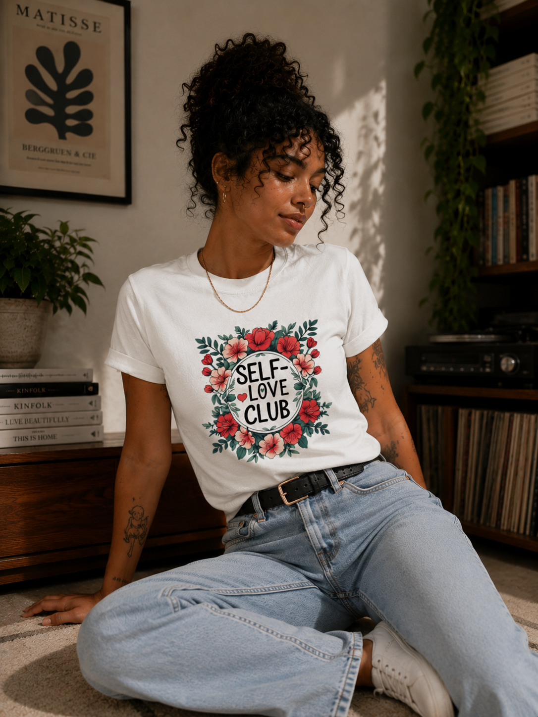A woman sitting on a carpet in a cozy room, wearing a white graphic t-shirt with a red and green floral design that reads 'SELF-LOVE CLUB'.