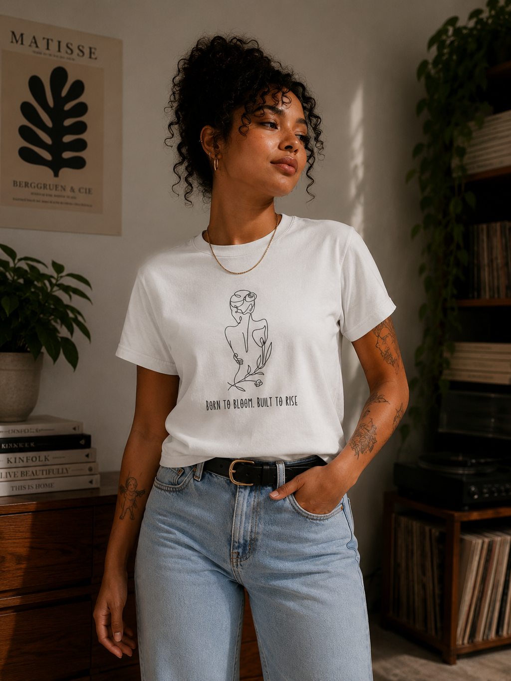 A woman wearing a white graphic t-shirt with a black line art illustration of a woman holding a flower and the text 'BORN TO BLOOM, BUILT TO RISE' paired with light wash blue jeans in a cozy, sunlit room.