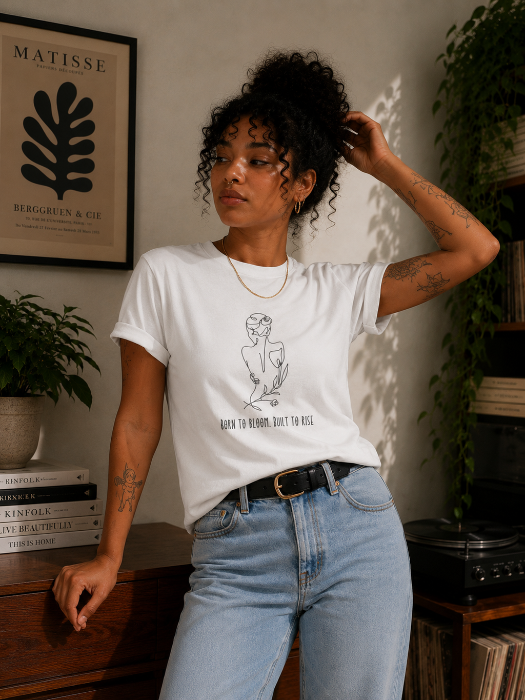 A woman wearing a white graphic t-shirt with a black line art illustration and the text 'I'M NOT A WOMAN, BUT I'M MIS'. She is posing in a cozy room with a wooden cabinet, plants, and a record player in the background.