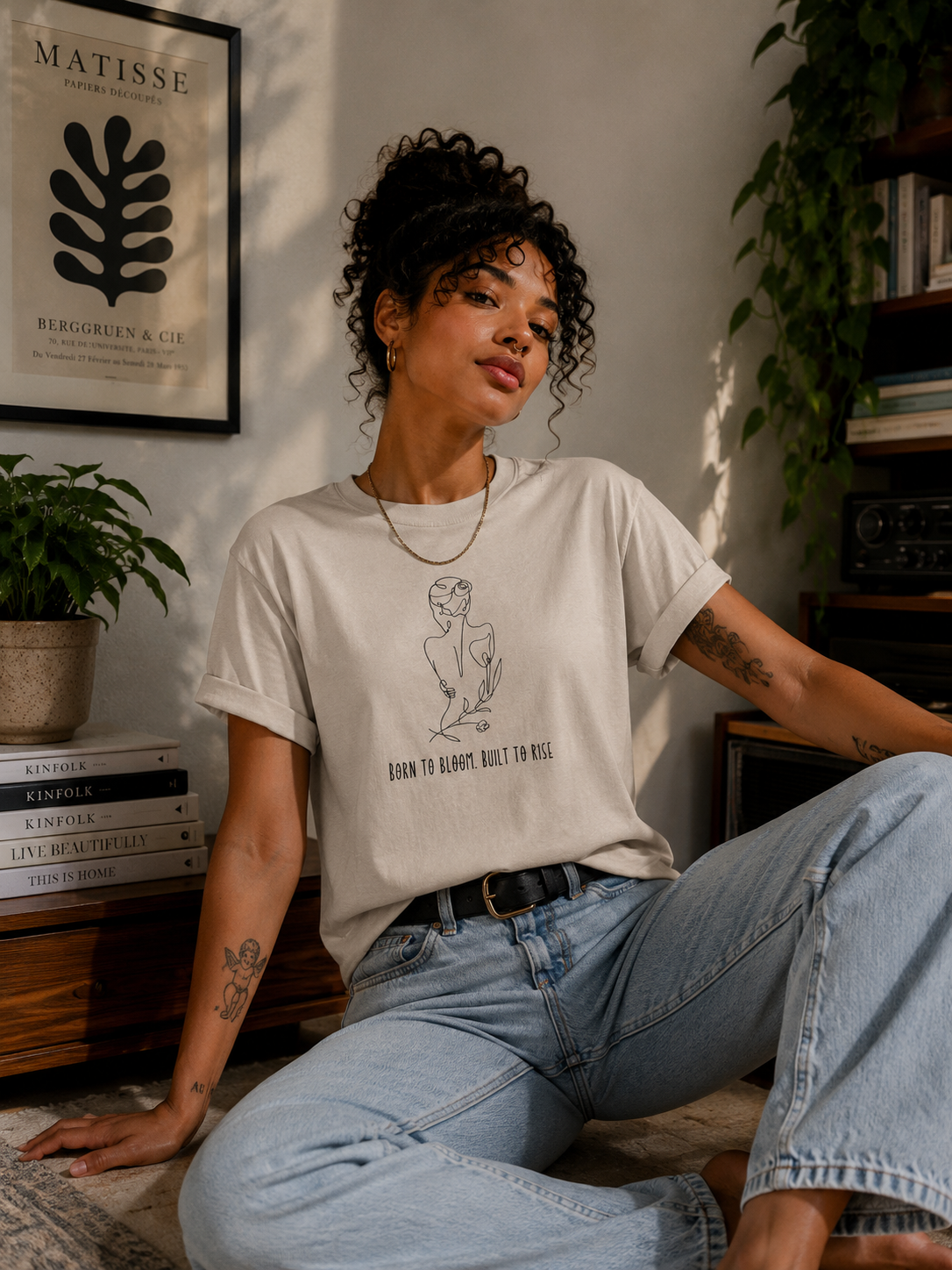 A woman sitting indoors wearing a light beige graphic t-shirt with a black line art illustration and the text 'BORN TO HAPPY, NOT TO FEAR'. She is also wearing light-wash blue jeans and a black belt, with a warm, sunlit room background containing books, plants, and framed wall art.