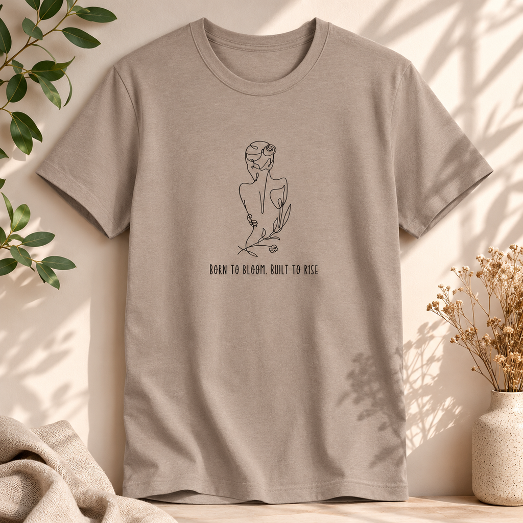 A taupe short-sleeve crewneck t-shirt with a black line art graphic of a woman in a flower crown and the text 'BORN TO BLOOM, BUILT TO KISS' on the front. The shirt is styled in a warm, sunlit setting with soft shadows, flanked by green leaves, a beige throw blanket, and a small vase of dried flowers.