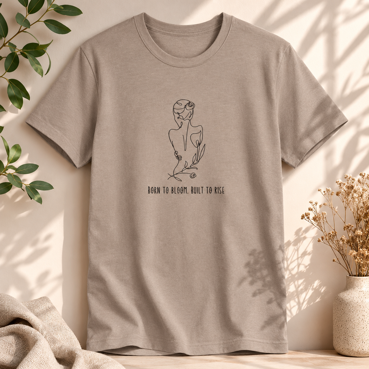 A taupe short-sleeve crewneck t-shirt with a black line art graphic of a woman in a flower crown and the text 'BORN TO BLOOM, BUILT TO KISS' on the front. The shirt is styled in a warm, sunlit setting with soft shadows, flanked by green leaves, a beige throw blanket, and a small vase of dried flowers.