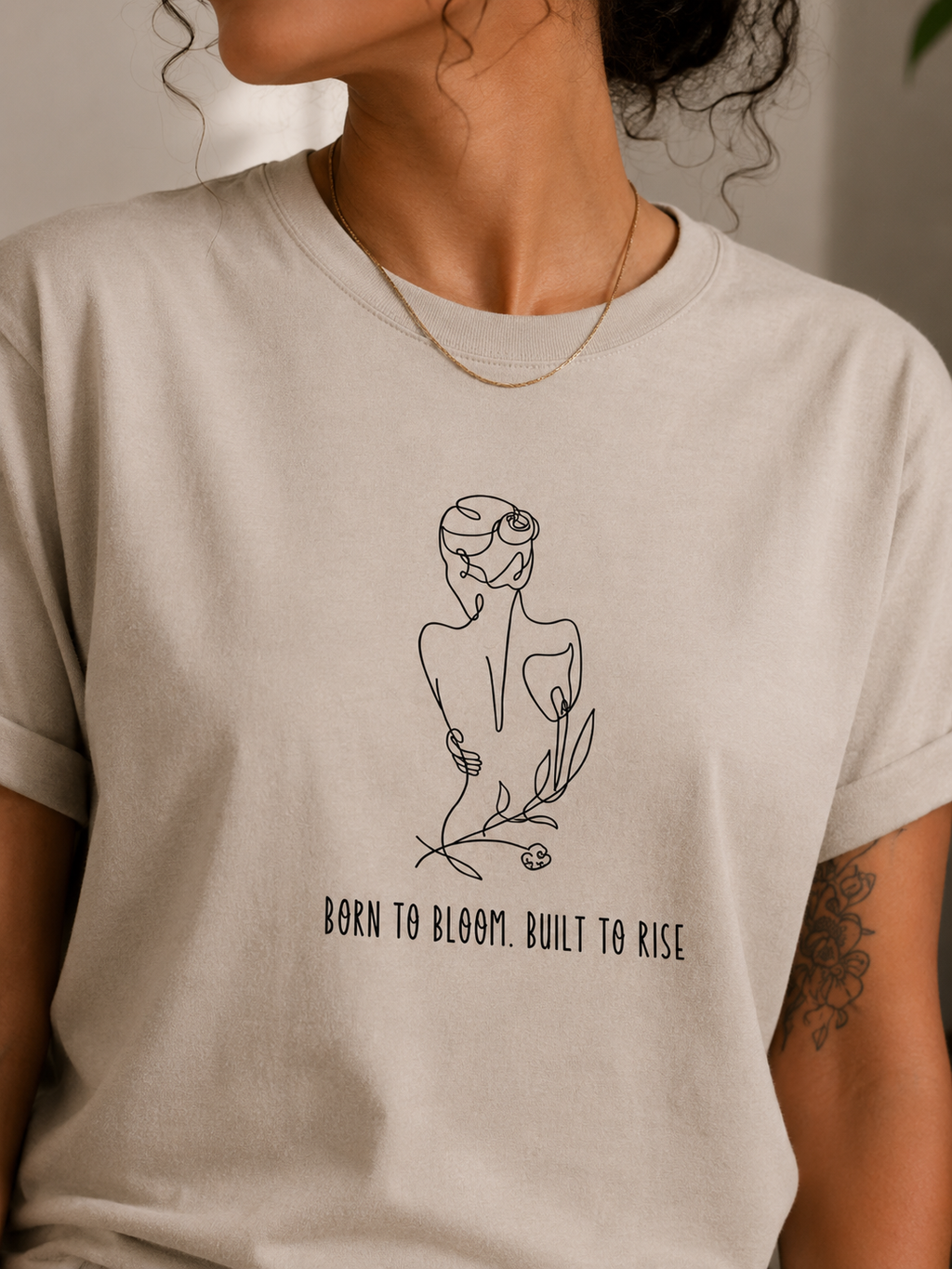 A close-up of a person wearing a light beige crewneck t-shirt with a black line art graphic of a woman in a flower crown and the text 'BORN TO BLOOM. BUILT TO RISE'.
