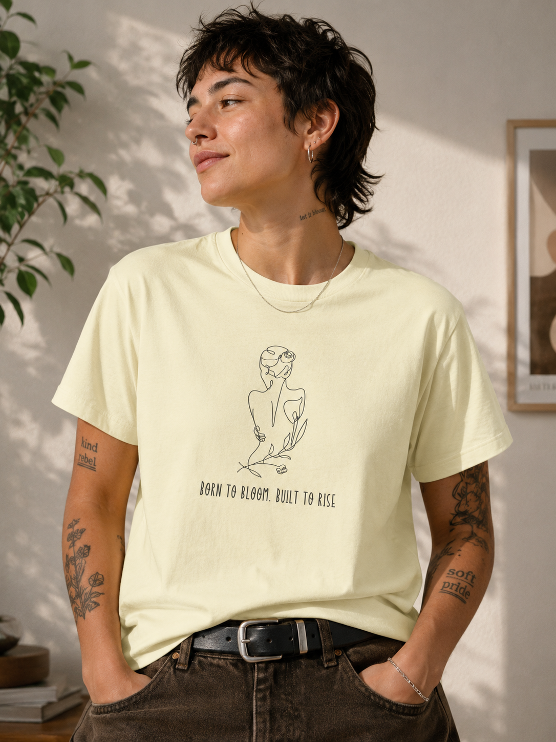 A person wearing a pale yellow graphic T-shirt with a simple line drawing of a seated figure and the text 'BORN TO BLOOM. BUILT TO RISE.