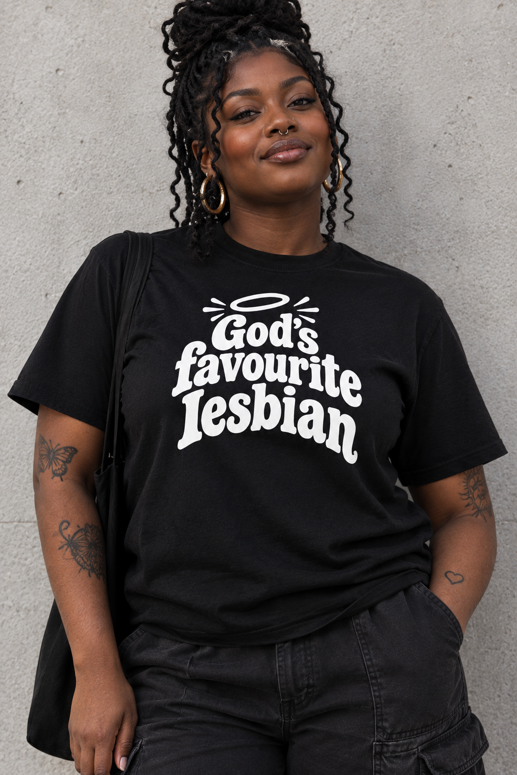 A woman wearing a black graphic t-shirt with the text 'God's favourite lesbian' in white stylized lettering, paired with black cargo pants, standing against a textured gray concrete wall.