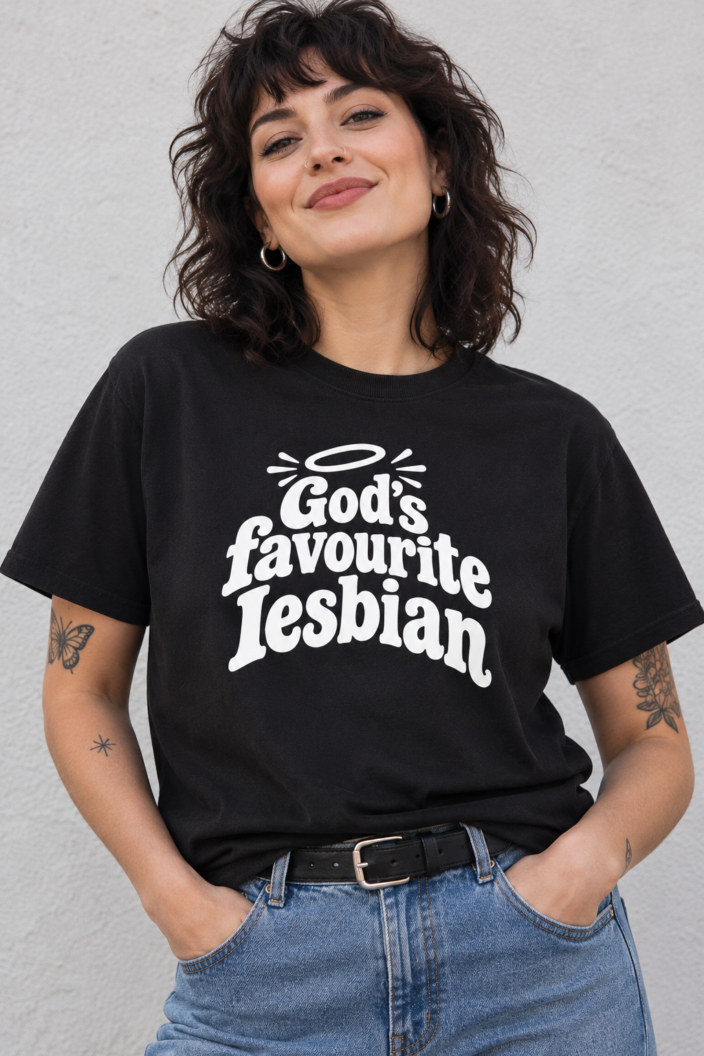 A woman wearing a black graphic t-shirt with the text 'God's favourite Lesbian' in white stylized lettering, paired with blue jeans and a black belt, standing against a light gray textured wall.